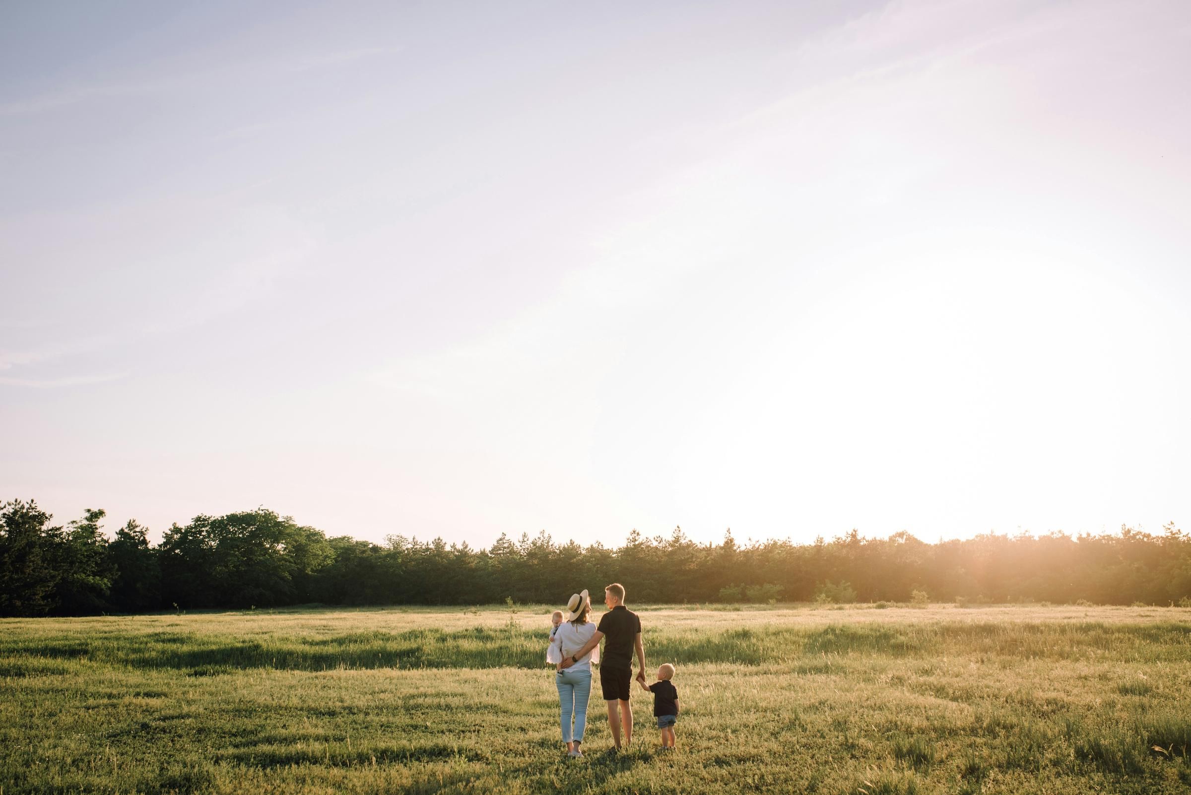 A family of three stands in a sunlit field, holding hands and facing the trees in the distance.