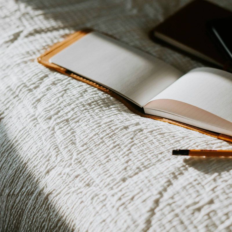 Open notebook, pencil, and another closed book resting on a textured white bedspread in sunlight.