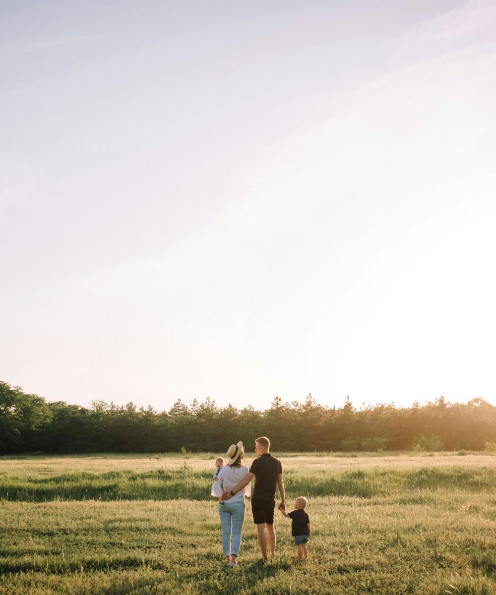 A family of three stands in a sunlit field, holding hands and facing the trees in the distance.