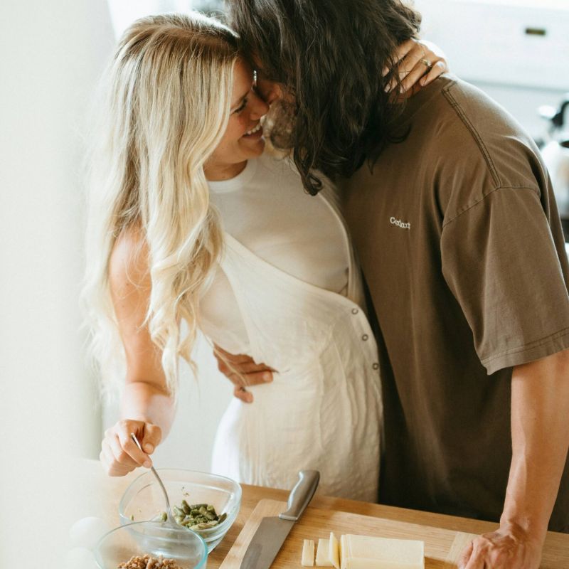 A couple embraces and smiles while preparing food together in a kitchen.